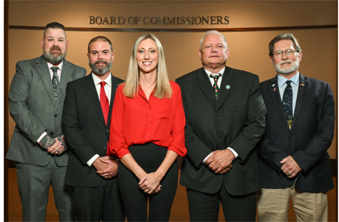 Group portrait of the five members of the Cabarrus County Board of Commissioners standing together in front of a wood-paneled wall beneath a Board of Commissioners sign.