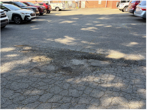 Photograph of a worn paved parking lot with cars parked in the background.