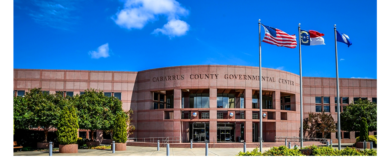 Photograph of the Cabarrus County Governmental Center, a curved brick building with U.S., North Carolina, and county flags flying in front under a blue sky.